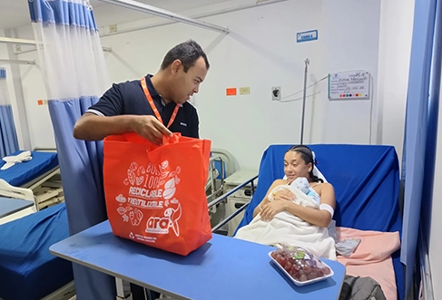 A woman with a newborn in a hospital bed gets a red bag full of groceries delivered by an Ara employee. (photo)