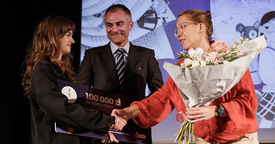 A person holding a 100,000zl check from Biedronka while shaking hands with a second person holding flowers. A smiling person is behind them (photo)