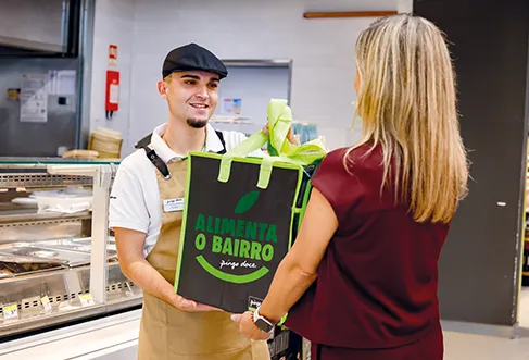 A Pingo Doce employee handing over a large reusable bag where its written "Alimenta o Bairro" to a customer infront of a counter (photo)