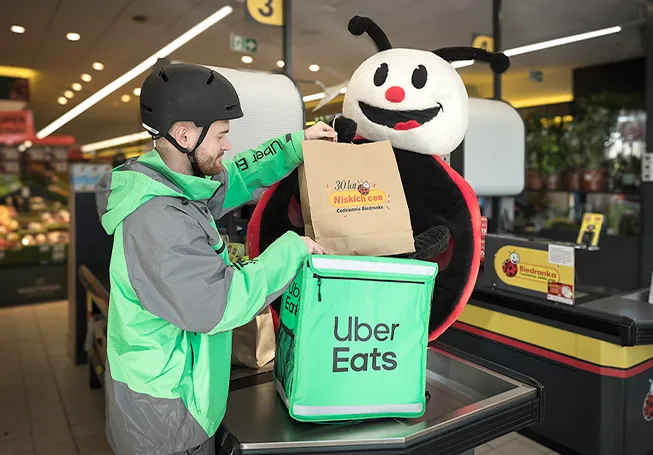 A Uber Eats delivery driver placing a paper bag into his bag at the check-out counter with a large Biedronka plushy behind him (photo)