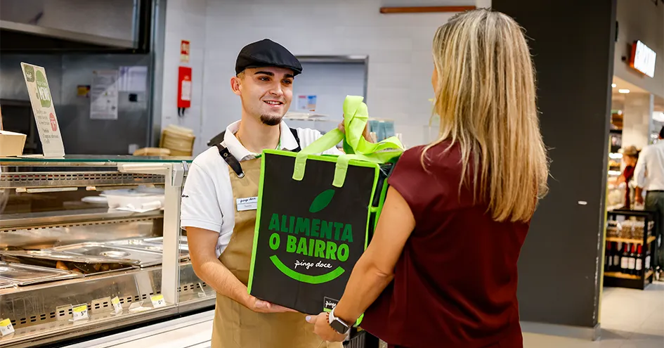 A Pingo Doce employee handing over a large reusable bag where its written "Alimenta o Bairro" to a customer infront of a counter (photo)