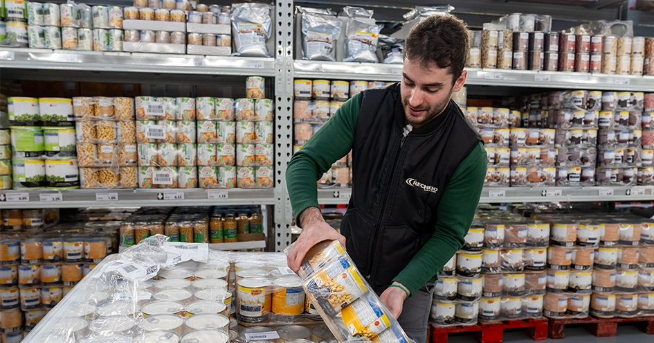 Um colaborador do Recheio está a repor latas de grão-de-bico em frente a uma prateleira que contém produtos enlatados (foto)