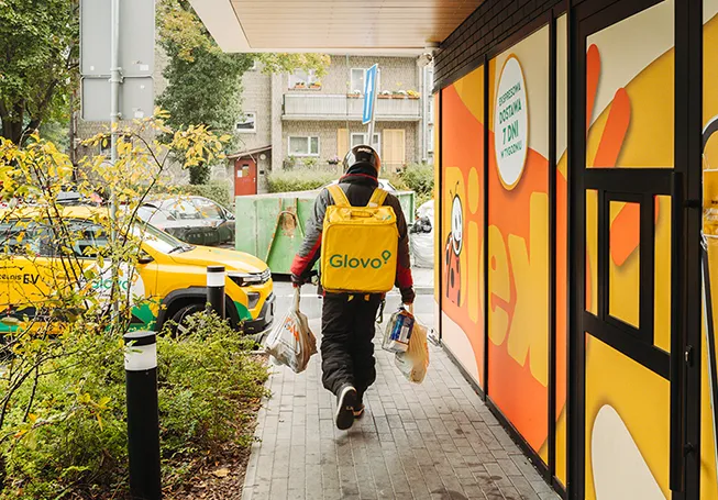 Um motorista de entregas com uma mochila Glovo a transportar vários sacos enquanto caminha ao longo de uma loja Biek, fotografado de costas (foto)
