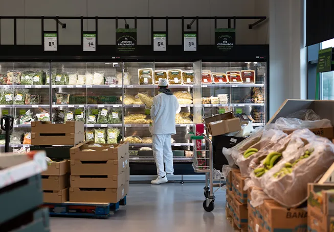 Colaborador fotografado de costas a reabastecer produtos hortícolas refrigerados, com caixas de produtos hortícolas em primeiro plano