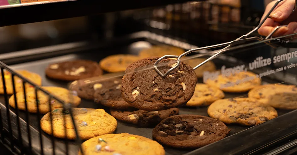 Grande plano de bolachas com pepitas de chocolate. Uma bolacha está a ser retirada com um par de pinças. (foto)