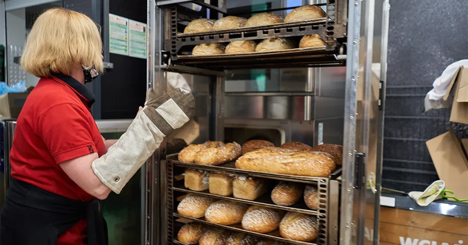 Colaboradora com luvas de proteção para forno a empurrar um carrinho com vários pães (foto)