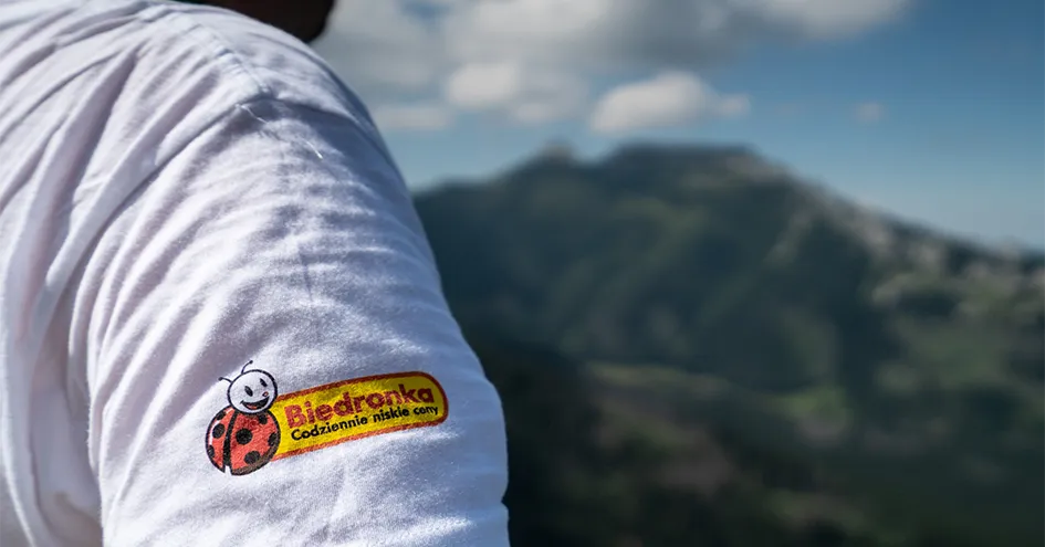Grande plano do ombro de uma pessoa, cuja t-shirt branca revela o logótipo da Biedronka, com uma montanha ao fundo. (foto)