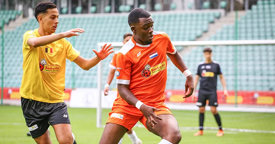 Dois jogadores adversários num jogo de futebol. Ambas as camisolas têm o logótipo da Biedronka. A baliza e o guarda-redes estão desfocados ao fundo.