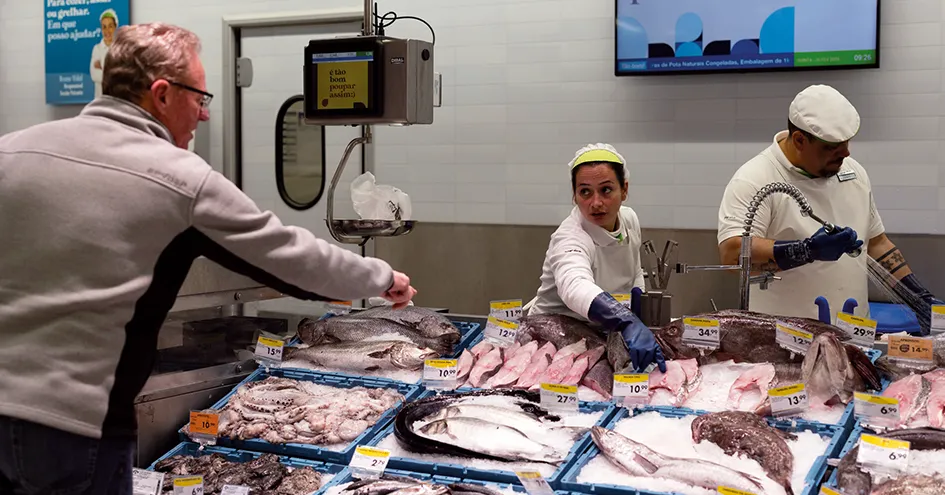 Uma variedade de peixe e marisco no gelo no balcão de marisco. Um cliente e um colaborador apontam para um peixe. Outro colaborador está a lavar um produto com uma mangueira, fora do enquadramento (foto)